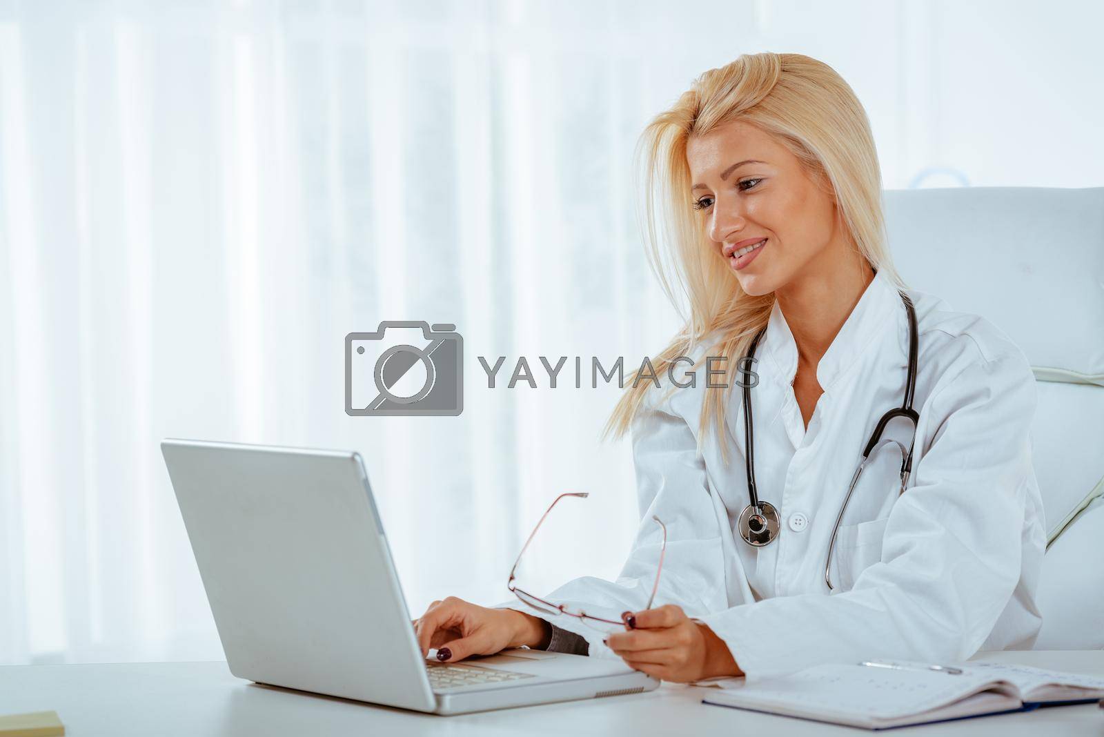Attractive blonde female doctor in her consulting room, sitting at the table in front of laptop and working.