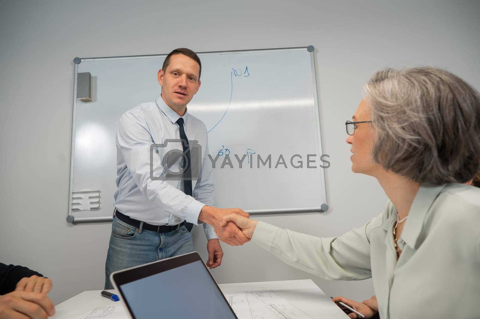 Royalty free image of The boss makes a presentation to subordinates at the white board. Caucasian man shaking hands with middle aged woman. by mrwed54