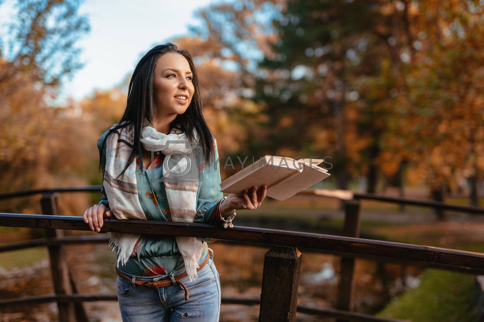 Young happy woman on a wooden bridge over the little lake in the park enjoying in beautiful autumn golden day with a book.