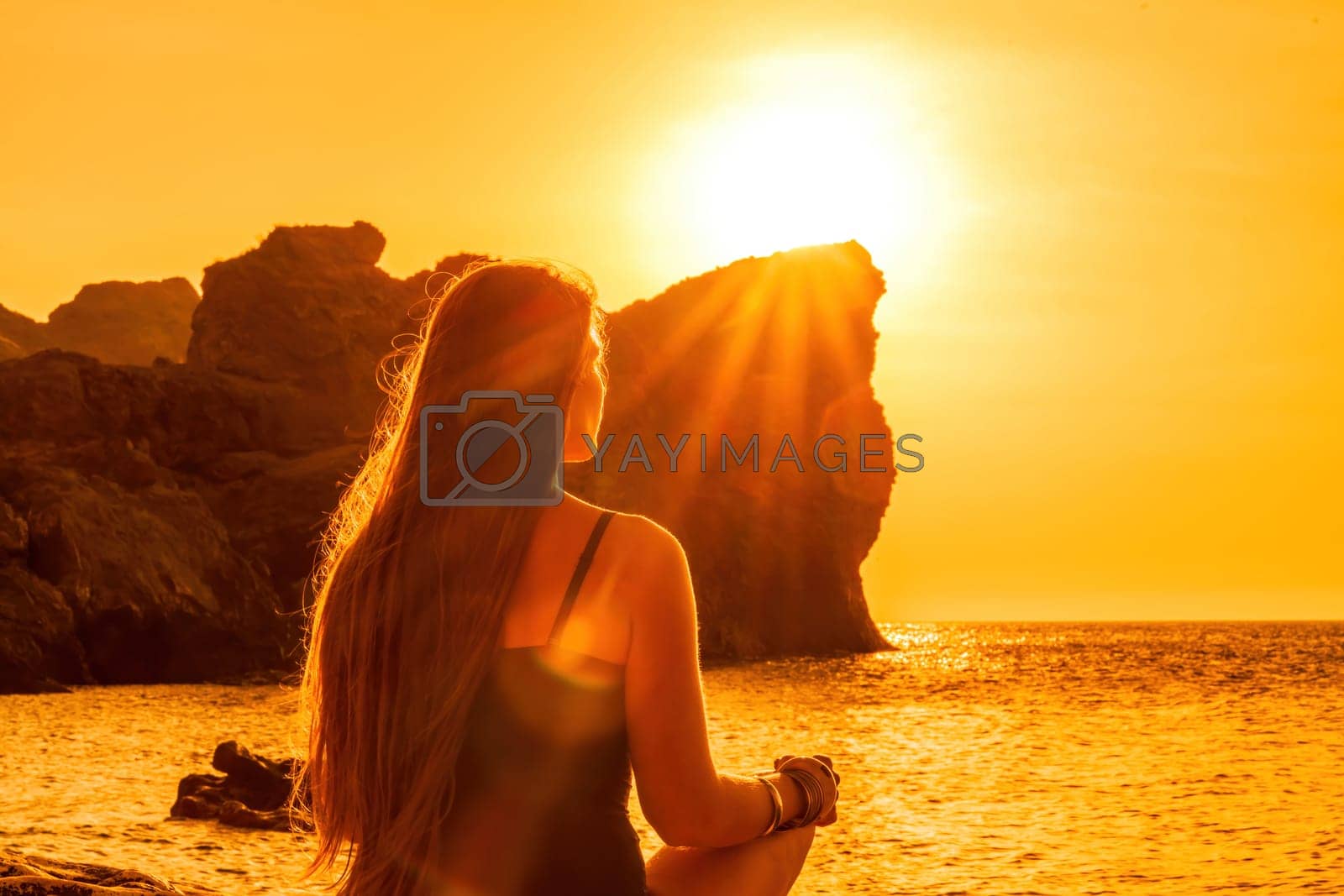Royalty free image of Woman tourist enjoying the sunset over the sea mountain landscape. Sits outdoors on a rock above the sea. She is wearing jeans and a blue hoodie. Healthy lifestyle, harmony and meditation by Matiunina