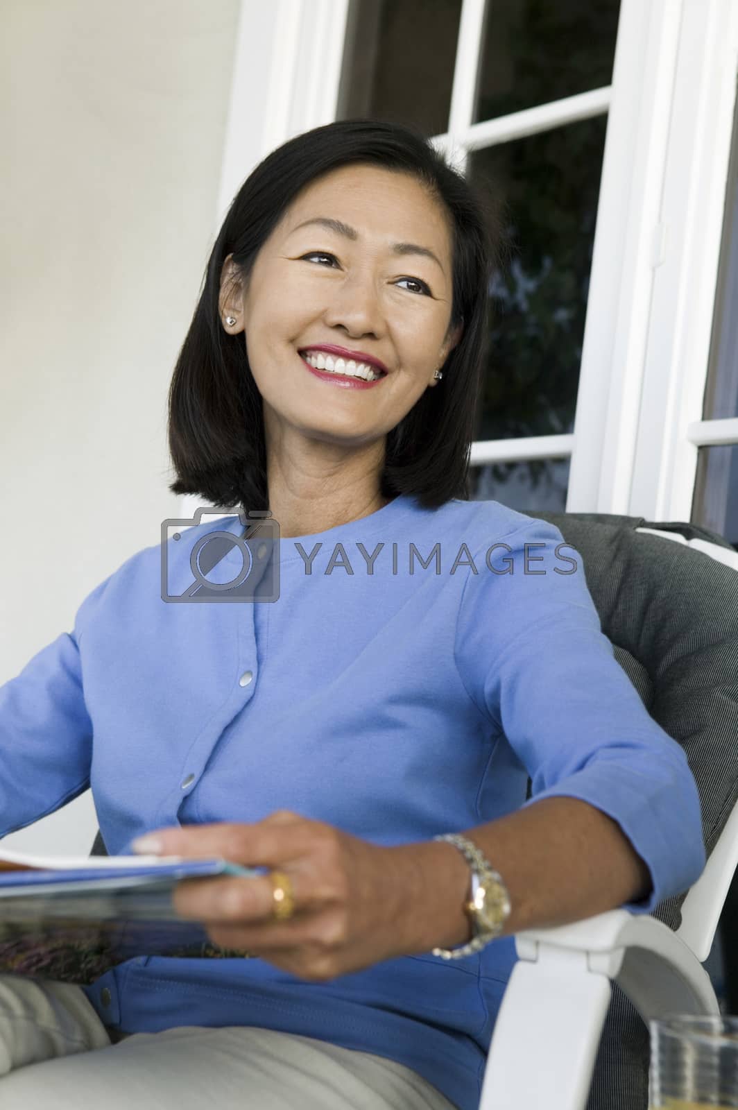 Royalty free image of Happy Asian woman looking away while sitting on chair holding magazine by moodboard