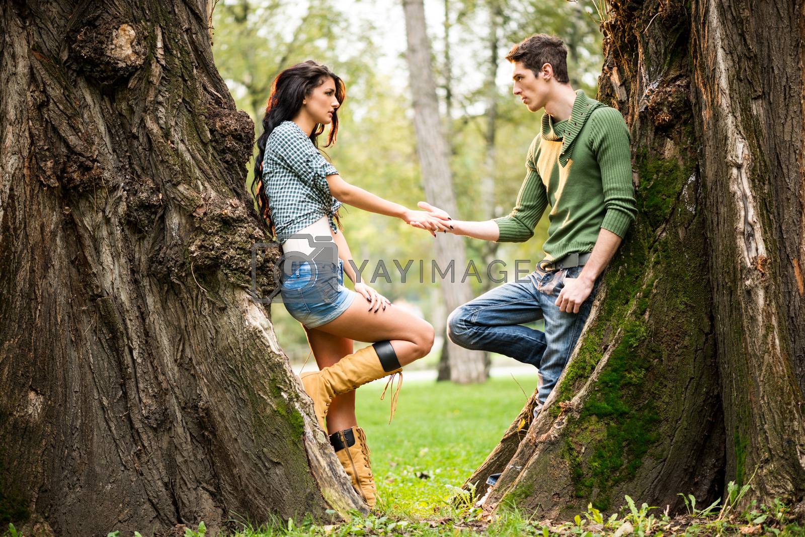 Young and pretty girl and guy at park leaning against a tree trunk, facing each other holding hands.