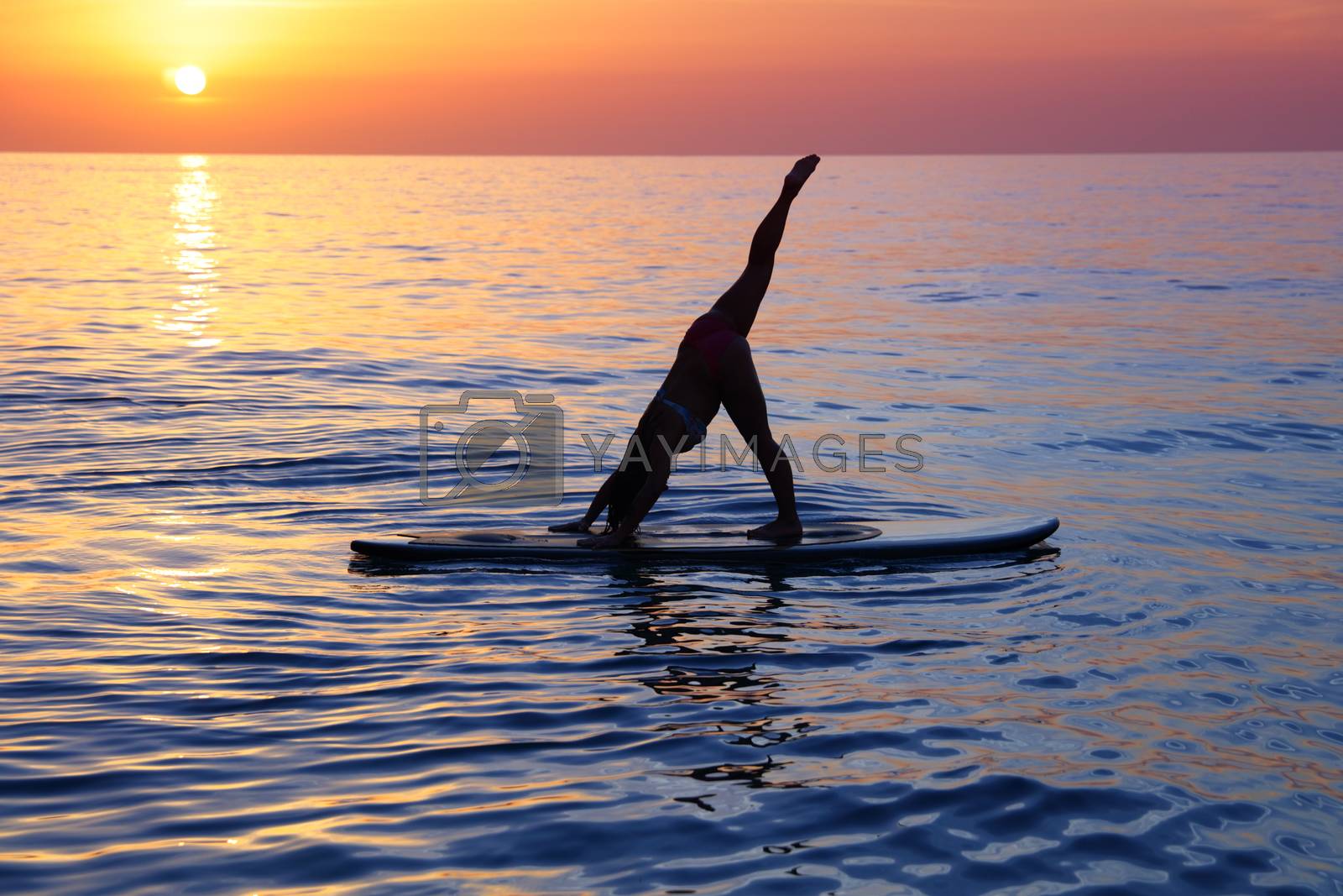 Sportive female doing yoga exercise on the beach over beautiful sunset background, standing on the sup board on the water in Pose dog muzzle down, Adho Mukha Svanasana
