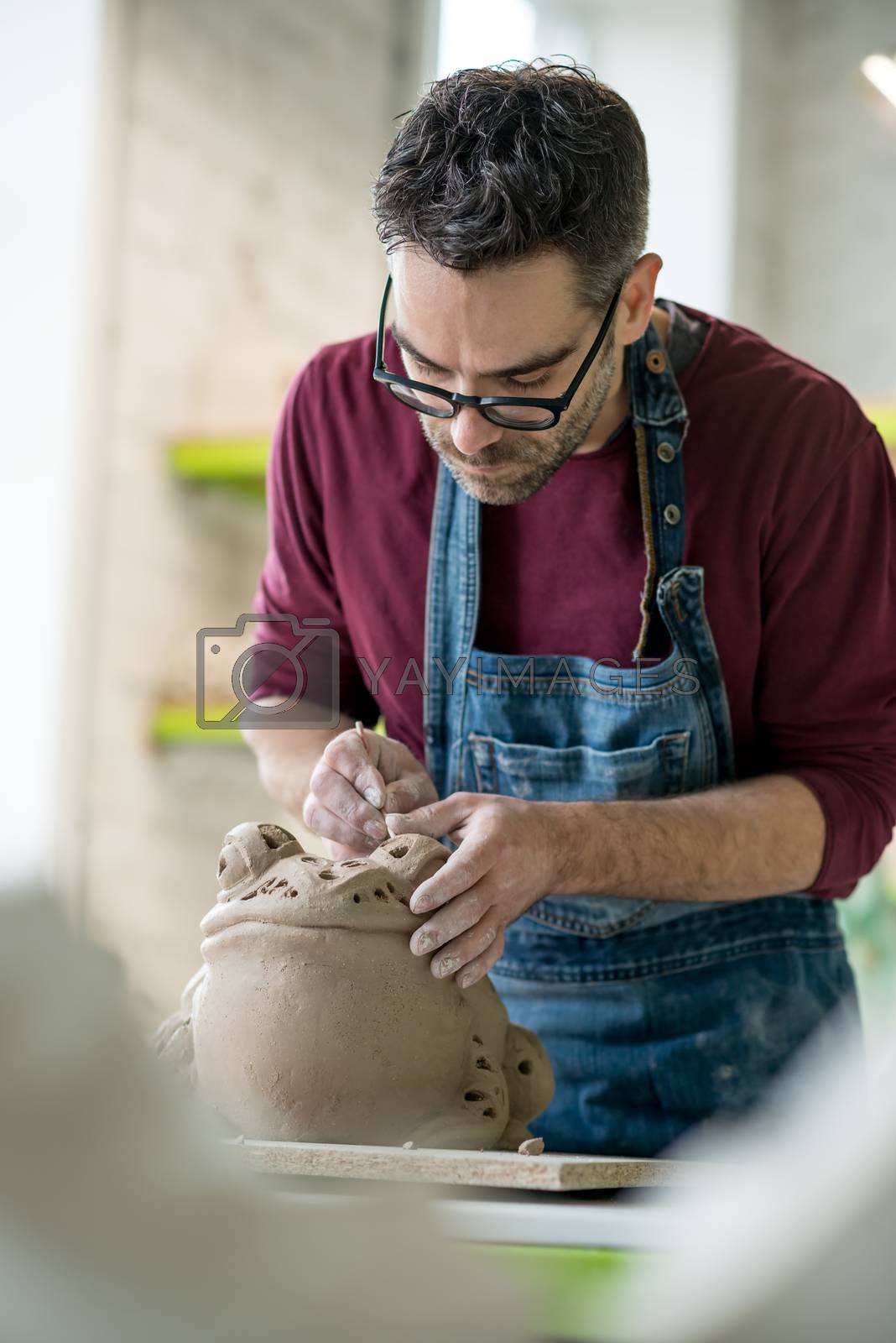Royalty free image of Ceramist Dressed in an Apron Sculpting Statue from Raw Clay in Bright Ceramic Workshop. by maxpro