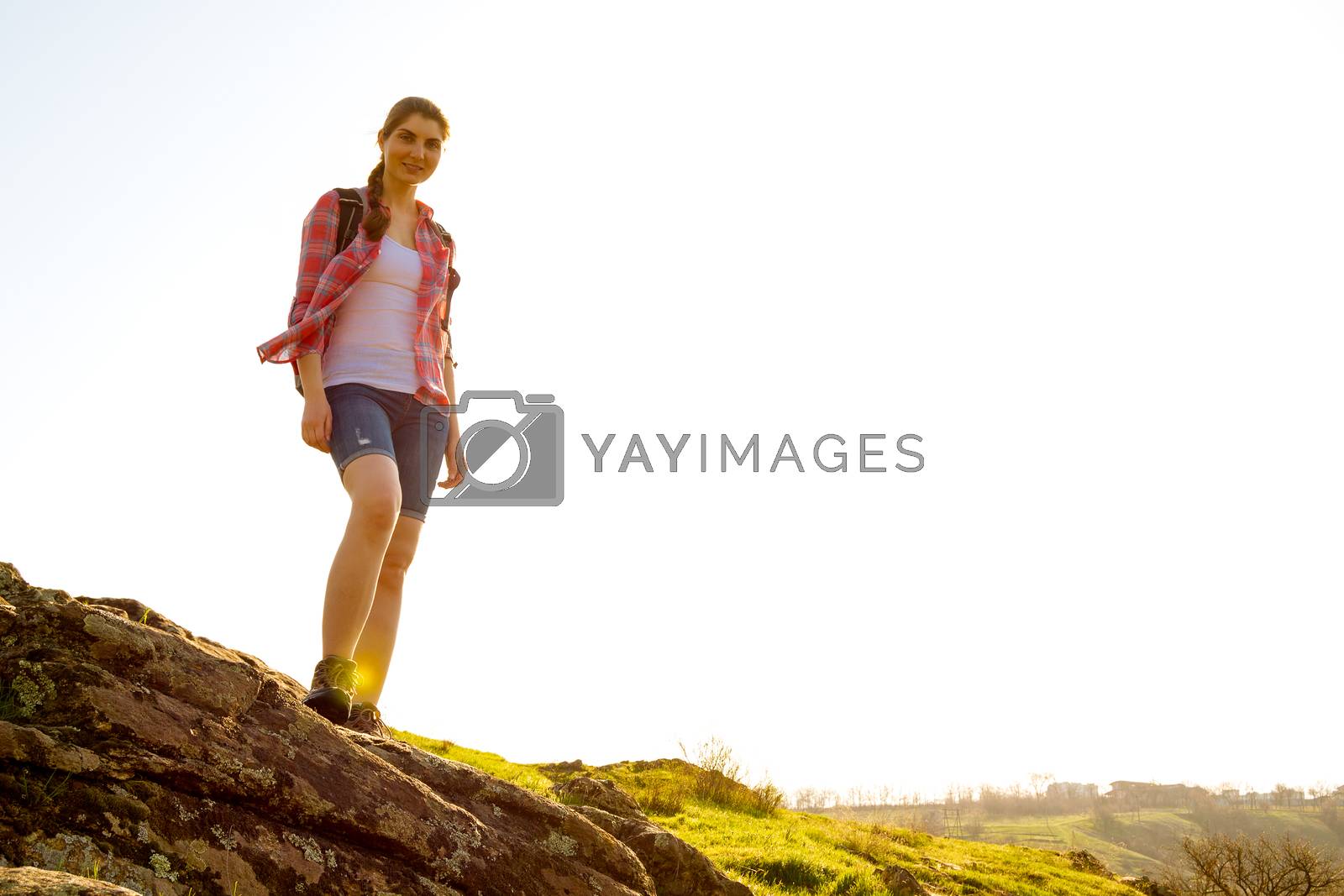 Royalty free image of Portrait of Happy Woman Traveler with Backpack Standing on the Rock at Sunny Evening. Travel and Adventure Concept. by maxpro