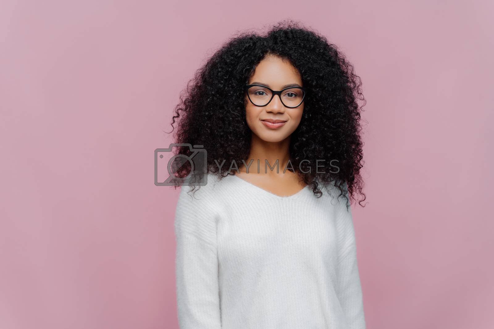 Royalty free image of Half length shot of attractive African American woman looks through transparent glasses, white sweater, has serious calm expression, poses against violet studio wall. Facial expressions concept by vkstock