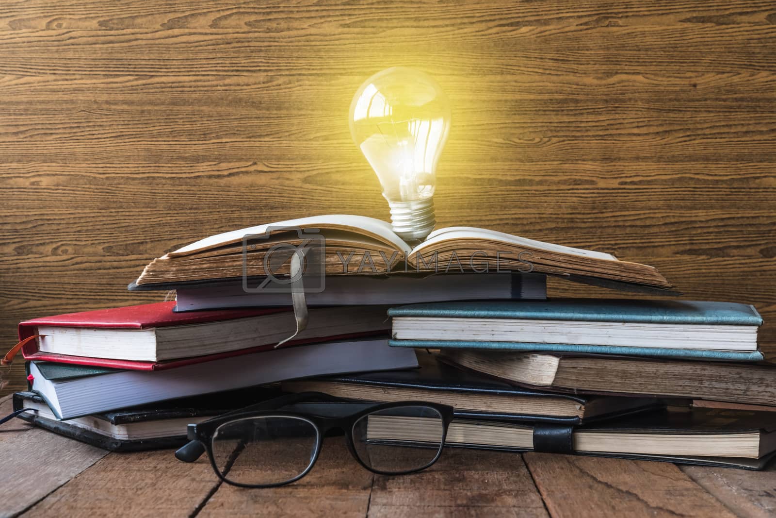 Royalty free image of Open book with light bulb, hardback books and glasses on wooden table. by ronnarong