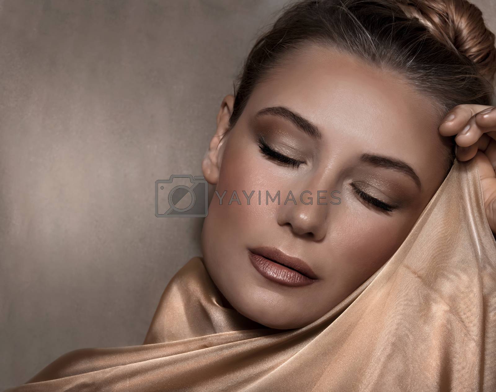 Closeup Portrait of a Beautiful Woman with Evening Makeup Wrapped in a Golden Silk Shawl over Beige Background. Luxury Fashion Look. 