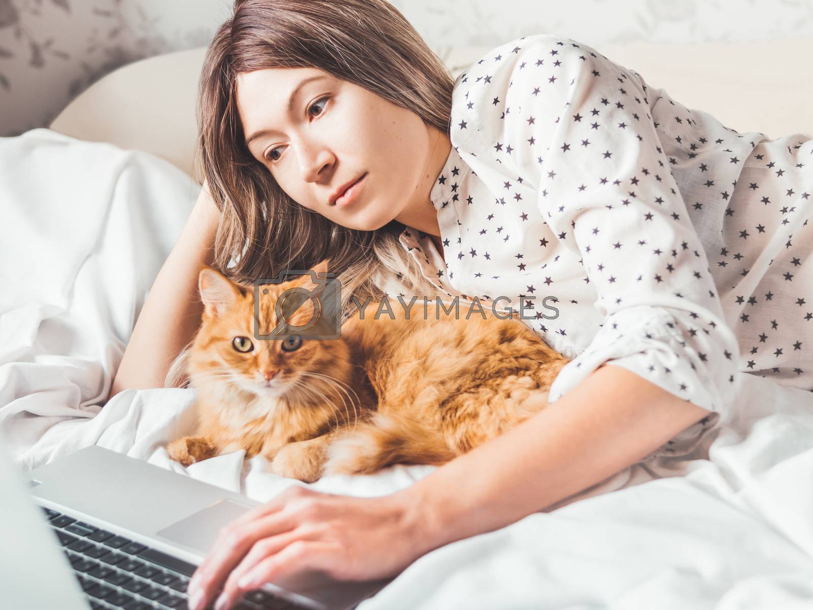 Royalty free image of Cute ginger cat and woman are lying in bed. Woman watching onlin by aksenovko