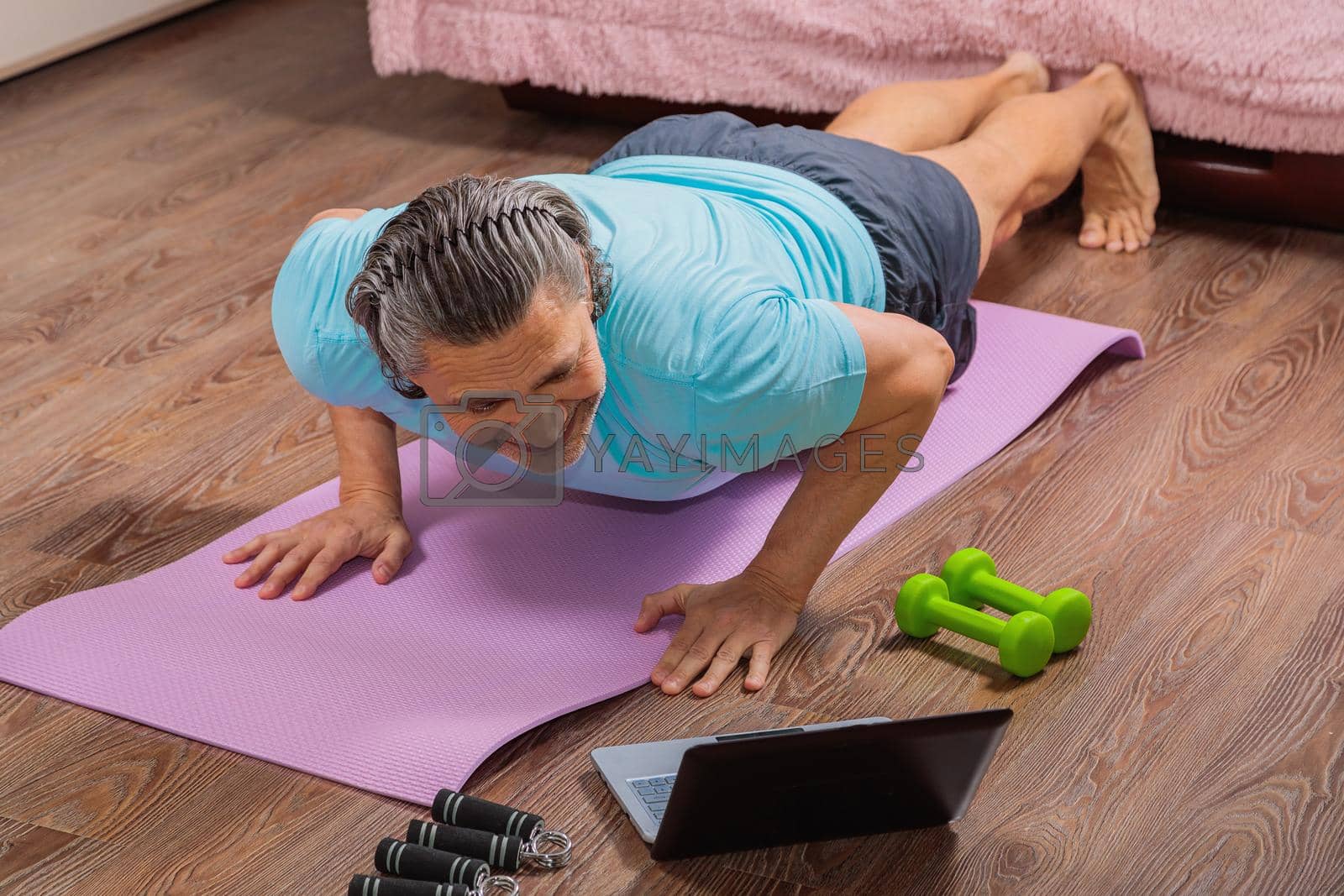 Royalty free image of 50 year old man performs exercises lying on mat at home looking at computer by Yurich32