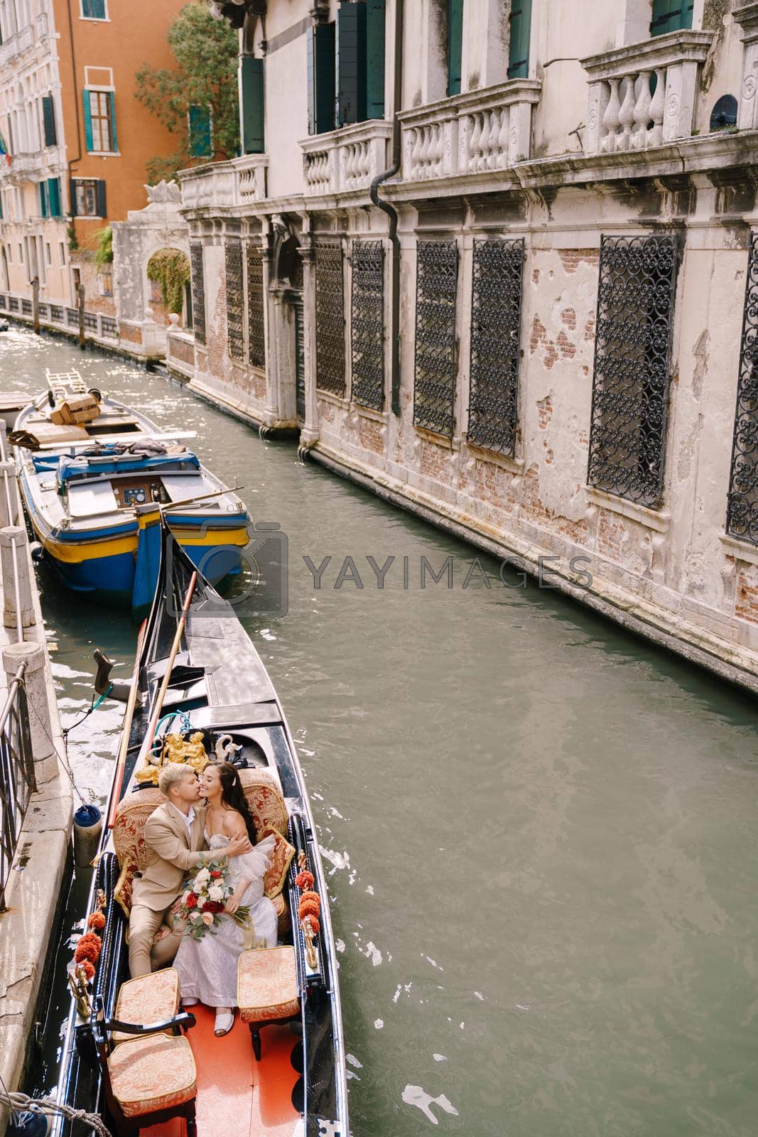 Royalty free image of Italy wedding in Venice. A gondolier rolls a bride and groom in a classic wooden gondola along a narrow Venetian canal. Newlyweds kiss in a boat moored to the shore. by Nadtochiy