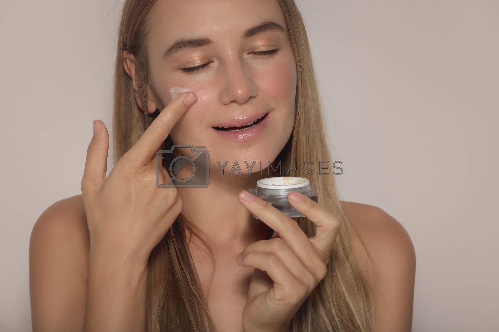 Portrait of a Beautiful Woman with Closed Eyes Applying Facial Moisturizer Cream. Natural Cosmetics. Isolated on Beige Background. Beauty Treatment. Healthy Lifestyle.