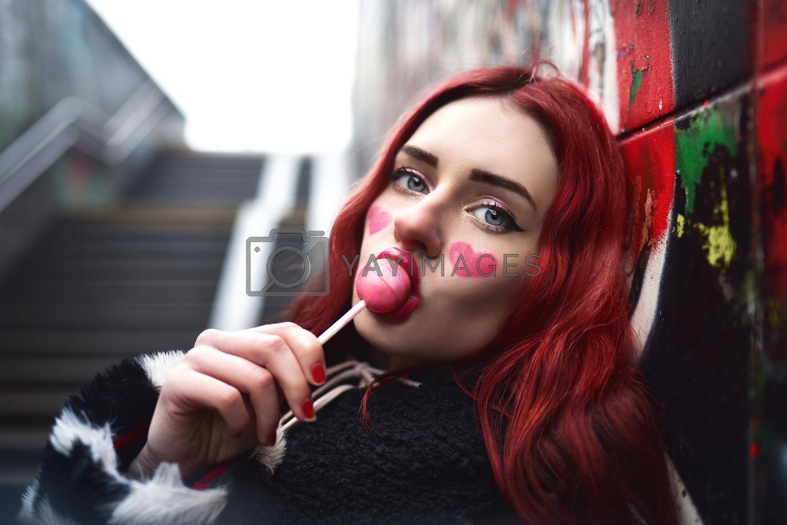 Royalty free image of A glamorous teenage girl with red hair licks a sweet strawberry candy near a wall of graffiti. by Nickstock