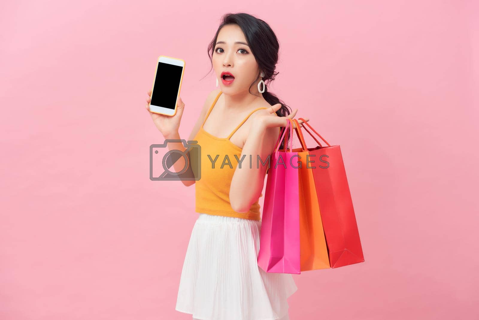 Royalty free image of Photo of shocked woman 20s in dress looking at smartphone in hand with surprise while holding shopping bags isolated over pink background by makidotvn