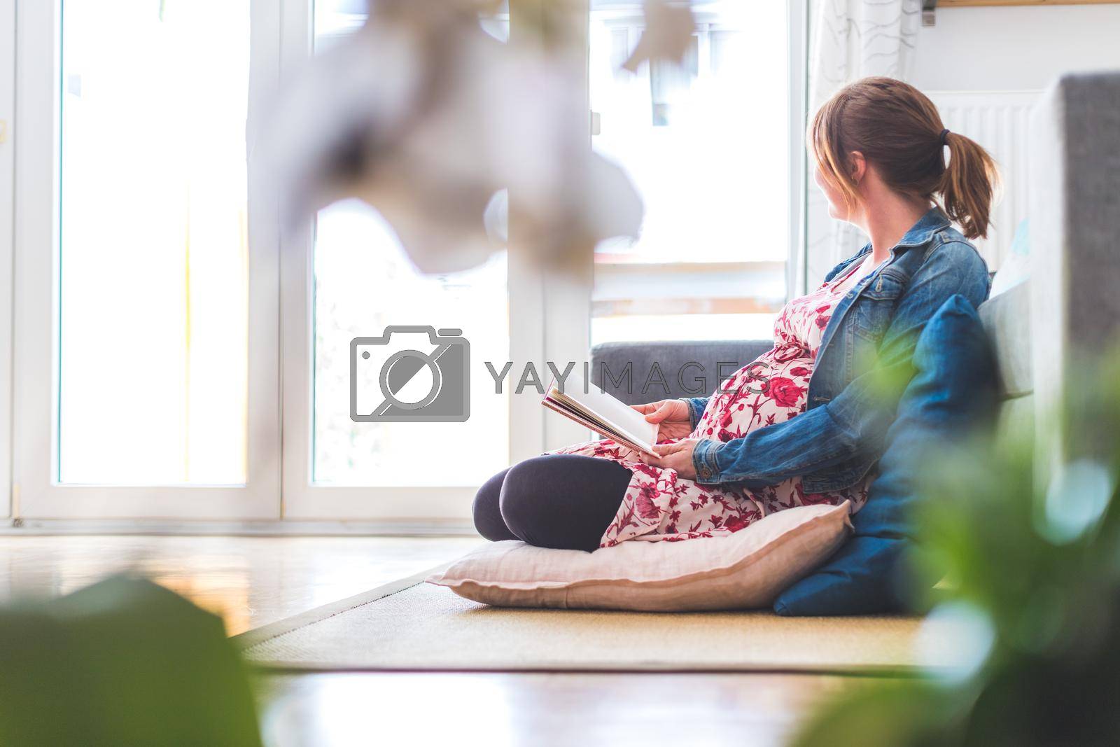 Pregnant caucasian happy mother is sitting on the floor, reading a book