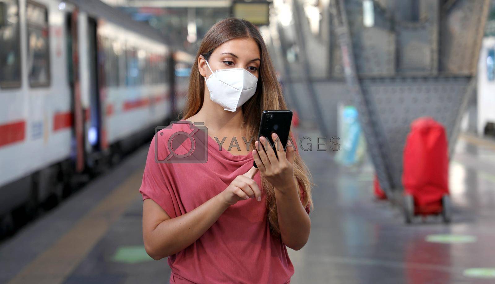 Young woman wearing KN95 FFP2 face mask waiting train at the station. Casual woman using smart phone in train station.