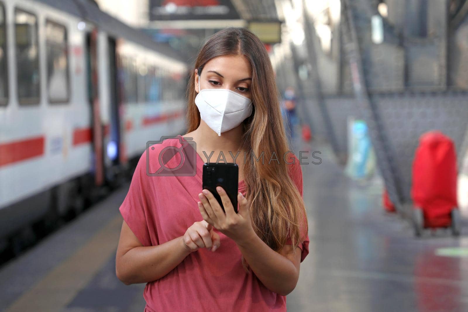 Young woman wearing KN95 FFP2 face mask waiting train at the station. Caucasian woman using smart phone in train station.