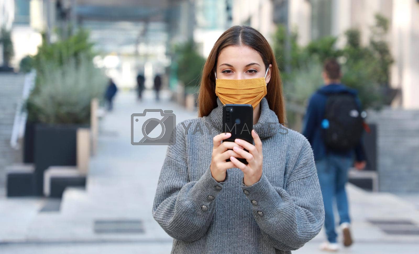 Royalty free image of Young woman with fabric mask messaging with mobile phone while walking in pedestrian street. Copy space. by sergio_monti