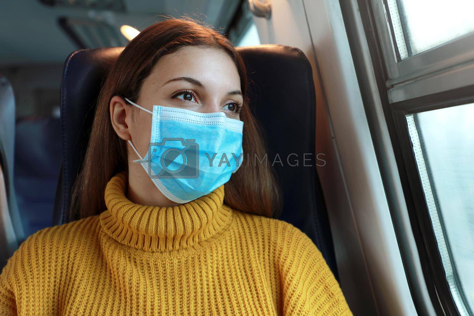 Close up of female commuter with surgical face mask looking through train window. Train passenger with protective mask travels sitting in business class looking through the window.