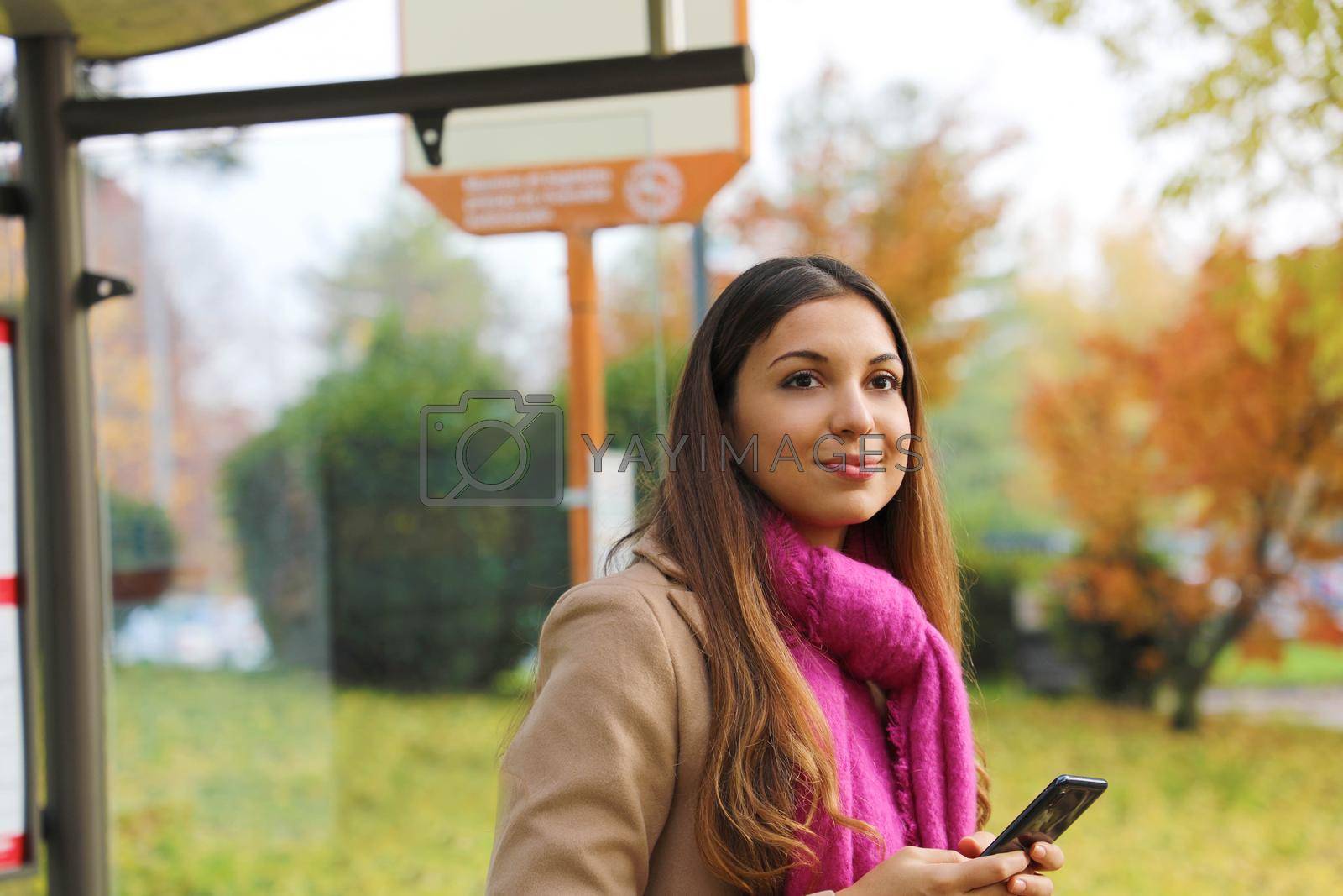 People and public transport. Beautiful young woman holding mobile phone waiting the bus on bus stop.