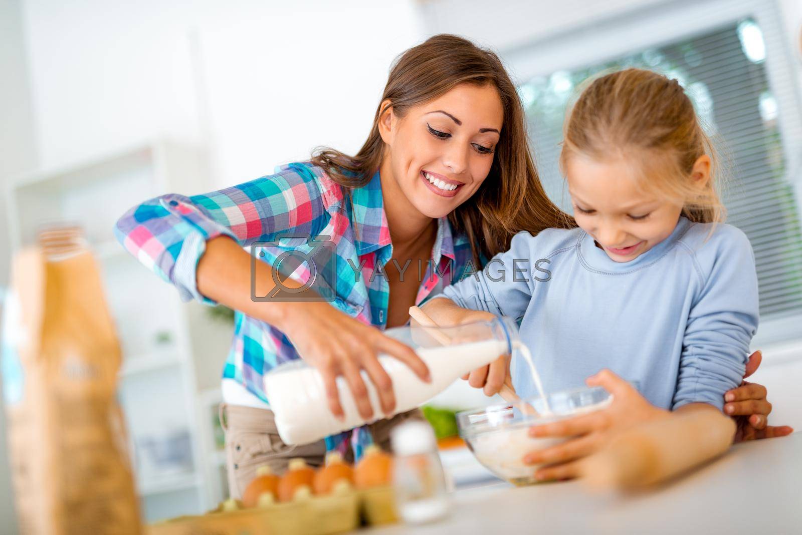 Mother and little girl are pouring the milk in the dish with dough in domestic kitchen. Mother learns her daughter how to make the cookies.