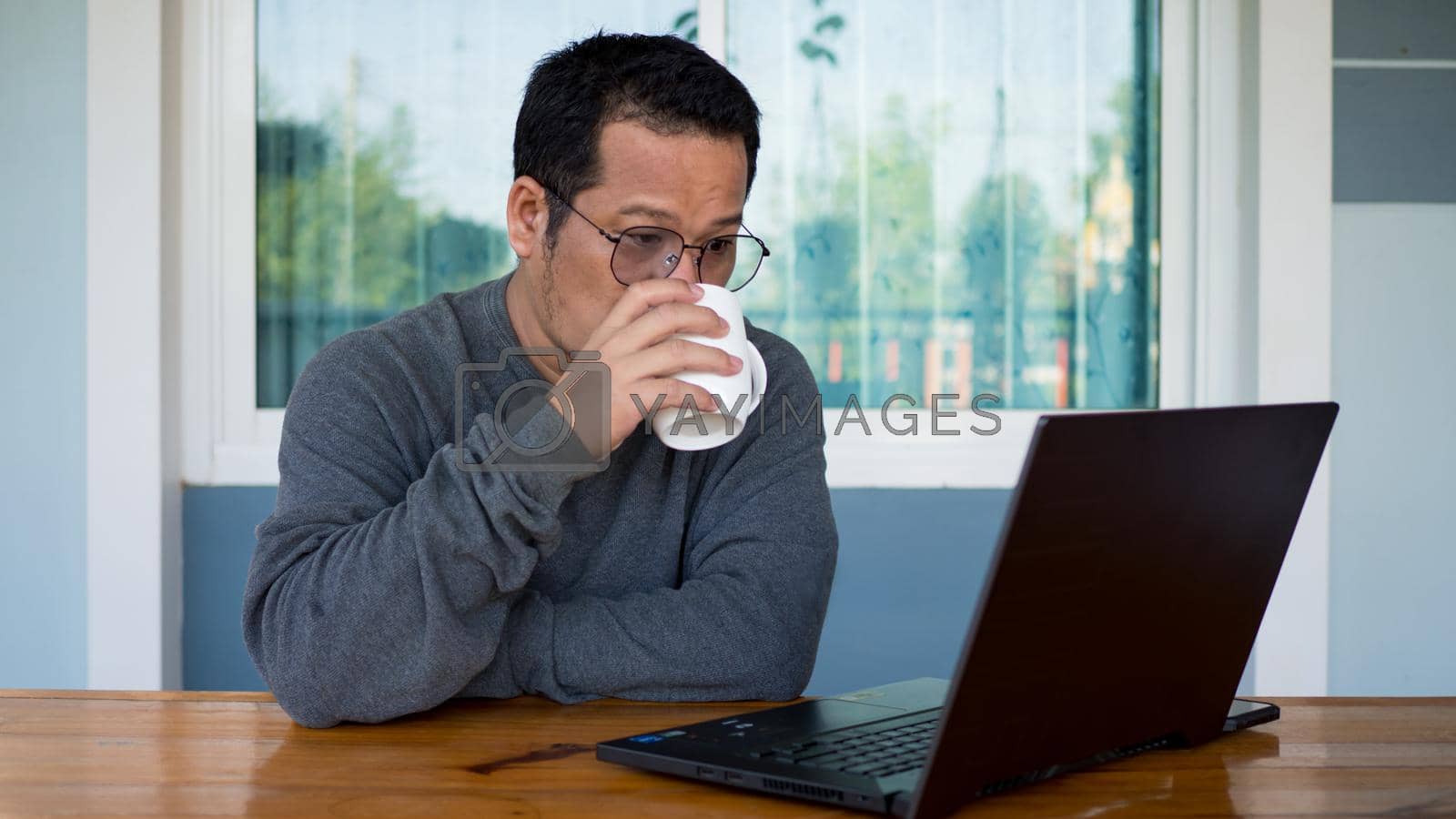 Royalty free image of Asian men wear long sleeves and eyeglasses.He is drink a coffee and Sitting at home working on a computer on a wooden table. Work from home concept. by Unimages2527