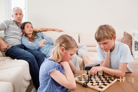 Siblings playing chess in front of their parents