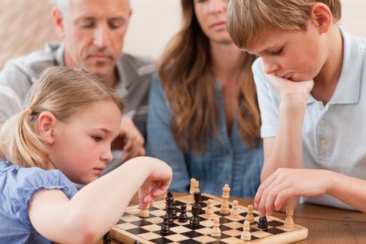 Focused siblings playing chess in front of their parents