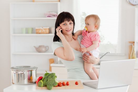 Charming brunette woman on the phone while holding her baby in her arms in the kitchen
