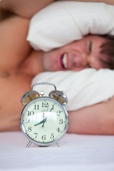 Angry young man lying in bed and annoyed by his alarm clock in t