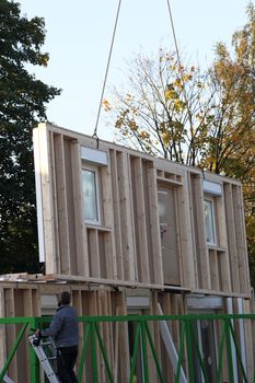 Crane hoisting up a prefabricated wall of a new build timber house from the trailer on which it has been delivered to the building site