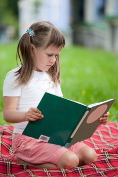 Little studious girl preschooler is reading open book and sitting on the red plaid on green grass in summer park