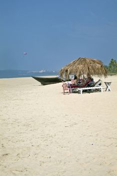 Portrait of secluded beach close of a city with day trippers and holidaymakers under the shade of coconut parasols. White sand, blue sky, sea, surf, para glider and fishing boats for a backdrop. Generic tropical shot location, Goa India
