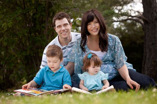Happy young family with pregnant mother sitting and reading in park