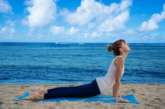 Young pretty woman practicing yoga on the beach by the ocean 