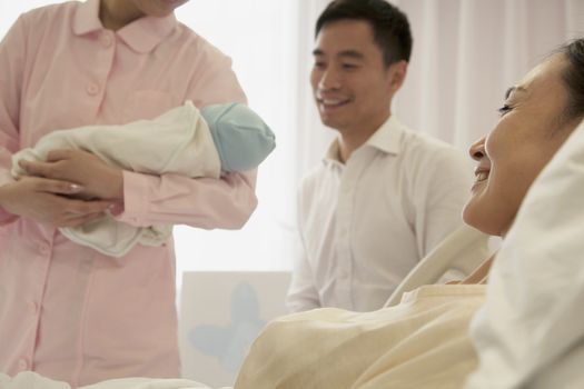 Nurse holding newborn baby in the hospital with mother lying on the bed and the father beside her
