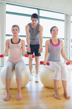 Two women at the gym with dumbbells sitting on exercise balls along with their coach