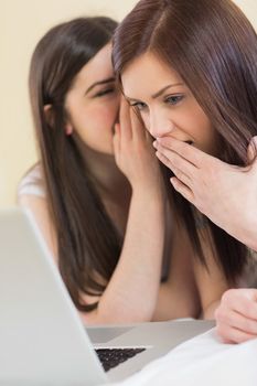 Pretty girl telling a secret to her friend in front of laptop at home in bedroom