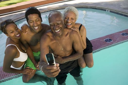 Senior couple and mid-adult couple posing for mobile phone photograph at swimming pool elevated view.
