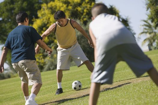 Boy (13-15) with two young men playing soccer outdoors in park.