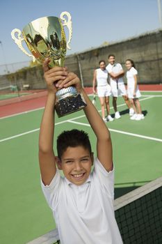 Portrait of a boy holding Tennis up trophy at net with adults in the background on tennis court