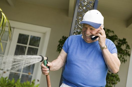Senior Caucasian man watering plants while enjoying a chat on cellphone