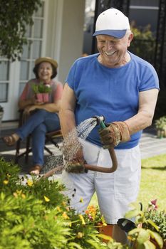 Happy Senior man watering plants in garden with woman sitting in background