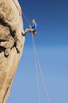 Low angle view of a young woman rappelling from cliff against clear blue sky