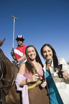 Portrait of happy female friends having champagne with polo player sitting on horse in the background
