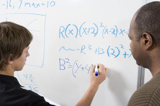 Young student writing math sums on board with professor standing on side