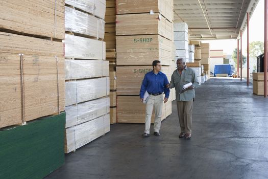 Two multiethnic men with clipboards walking along stacks of wood in warehouse