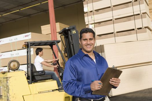 Portrait of a male supervisor with clipboard and forklift truck driver in the background