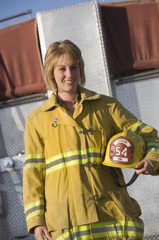 Portrait of a happy middle aged female firefighter holding helmet with fire brigade in the background