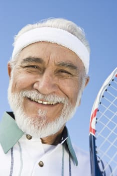 Close-up portrait of a happy senior man holding tennis racquet against sky